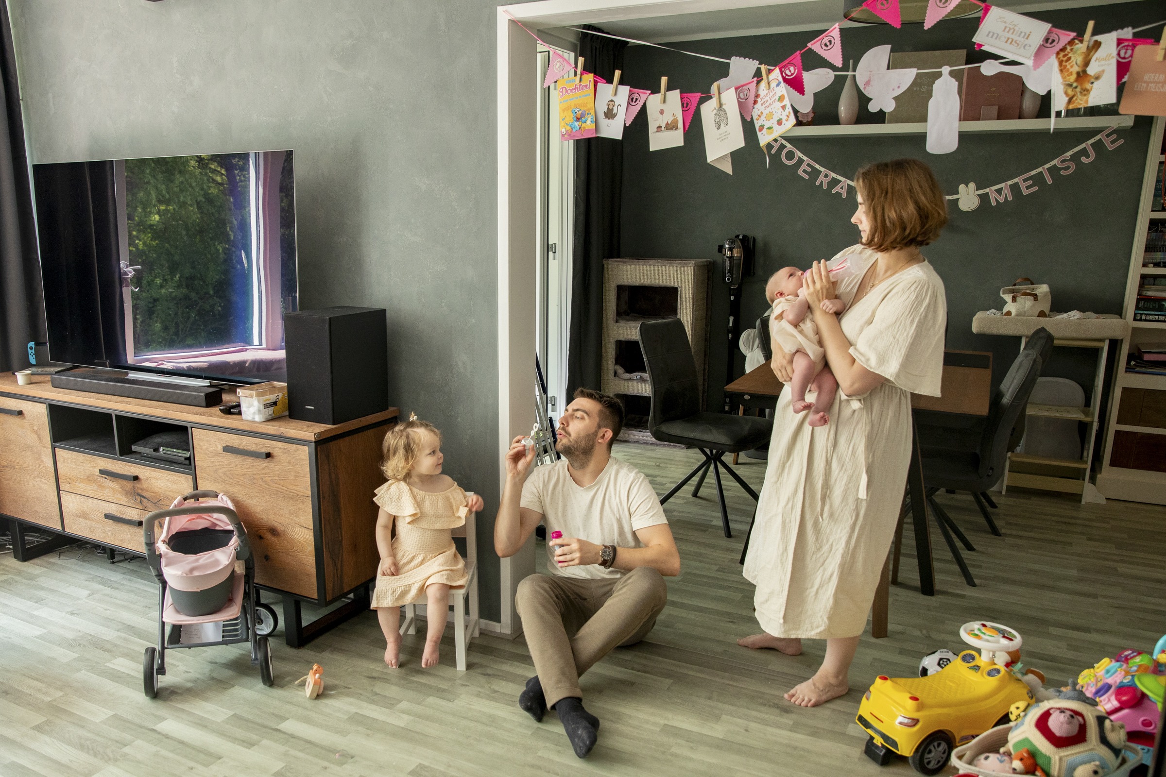 Gezin in de woonkamer, moeder heeft baby op de arm en vader is bellen aan het blazen terwijl dochtertje toe kijkt op haar stoeltje